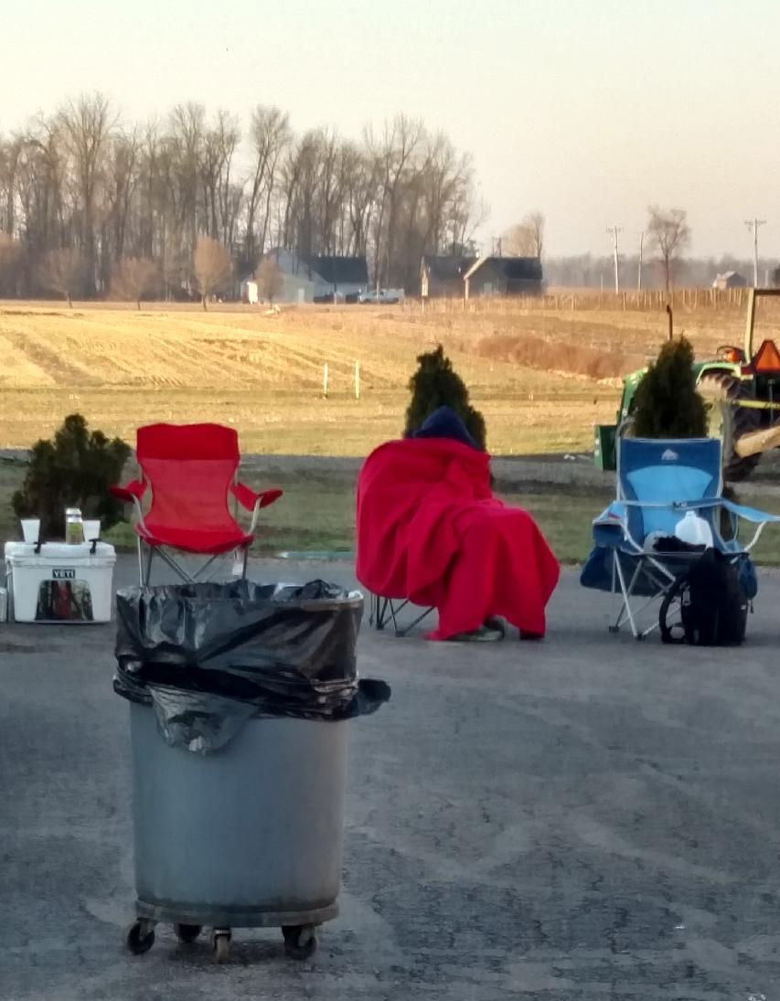 Hard two-tone duotone photograph of empty red and blue camp chairs, a trash bin and a cooler on pavement beside open fields and trees, quiet and expectant festival mood