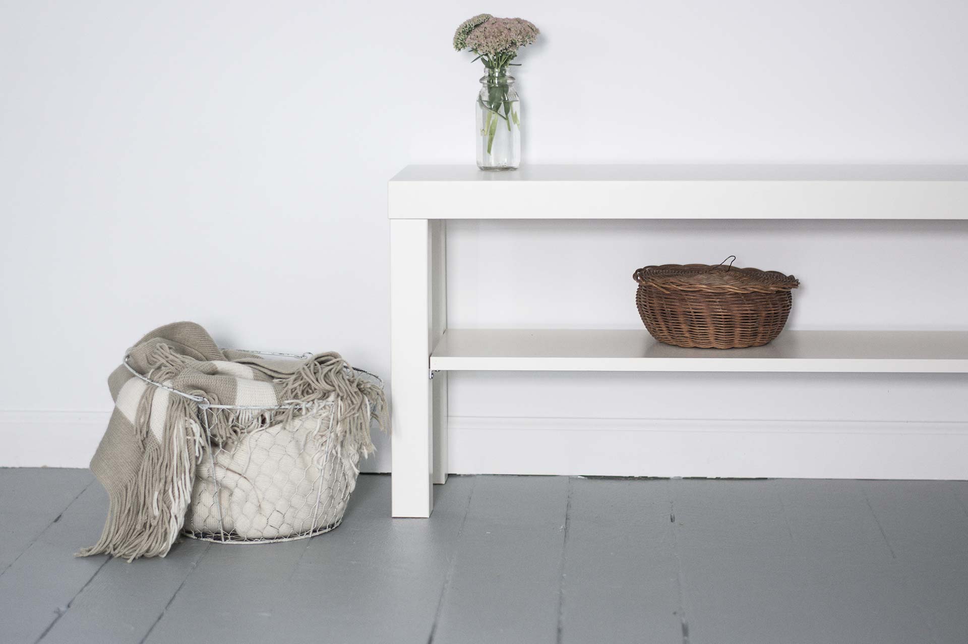 Soft black and warm gold duotone photograph of a white console table against a white wall, holding a glass vase with flowers, a wicker basket underneath, and a wire basket on top, calm and inviting lifestyle mood